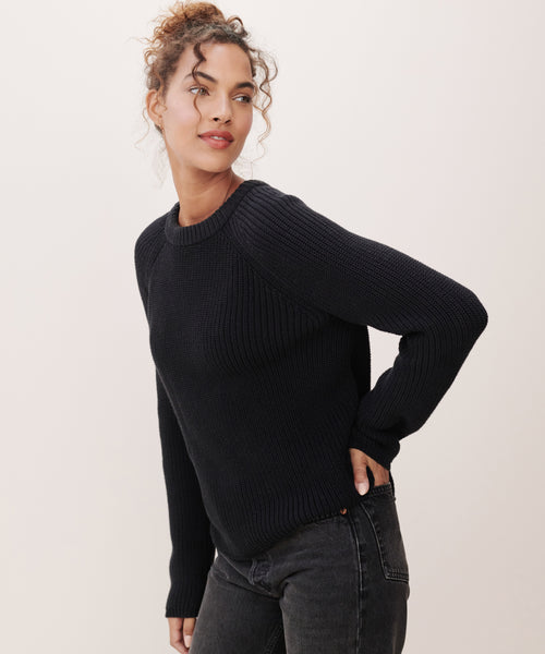 A woman with curly hair wears the Cotton Fisherman Sweater in black and black jeans, standing against a light background and gazing to the side with a relaxed expression.