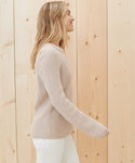 A woman with long blonde hair smiles while standing sideways in front of a light wooden wall, wearing the Cotton Fisherman Sweater in light beige and white pants.