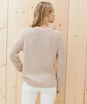 A woman with blonde hair, seen from the back, wears the Cotton Fisherman Sweater in light beige and white pants, standing before a light wooden wall.