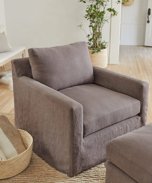A modern living room featuring a plush, charcoal armchair in Fabric Swatch Charcoal Linen on a woven rug, a matching ottoman, a basket with blankets, a potted plant, and light wood floors.