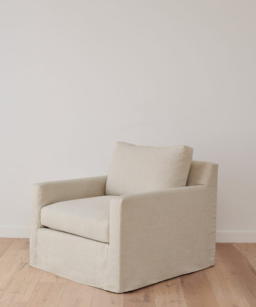 The Harbor Chair, upholstered in beige natural linen and handmade in Los Angeles, features a cushioned seat and backrest. It is placed on a light wood floor before a plain white wall.