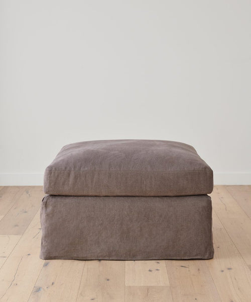 A brown, square Harbor Ottoman with a soft cushion from Jenni Kayne Furniture rests on a light wood floor against a plain white wall.