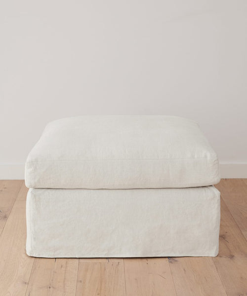 The Harbor Ottoman, handcrafted in Los Angeles, showcases soft edges and ivory linen upholstery, elegantly placed on a light wood floor with a simple white wall as the backdrop.