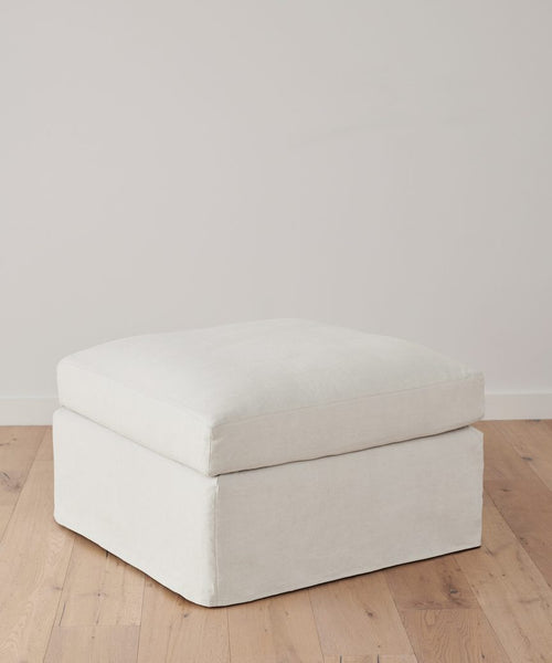 A Harbor Ottoman, crafted in Los Angeles with clean lines, sits on a light wooden floor against a plain, pale wall.