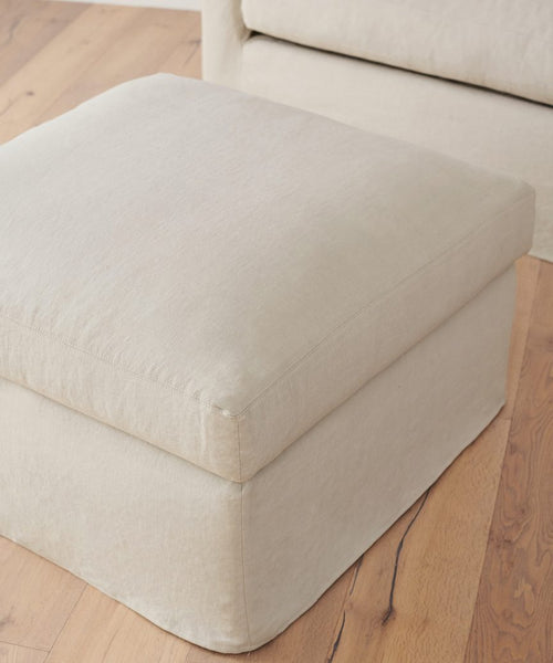 A handmade Harbor Ottoman in natural linen, beige and square, is placed on a wooden floor near a matching armchair in Los Angeles.