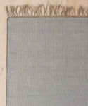 A close-up of the Hudson Jute Rug, handwoven in light gray with fringed edges, shown partially on a light wooden floor. Only the upper portion and left side of this natural fiber rug are visible.