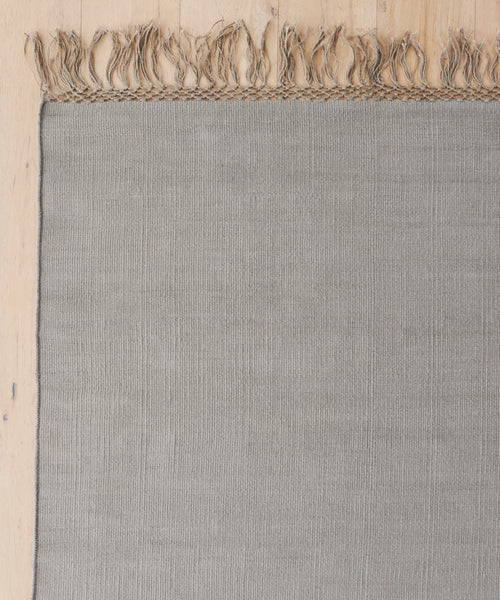 A close-up of the Hudson Jute Rug, handwoven in light gray with fringed edges, shown partially on a light wooden floor. Only the upper portion and left side of this natural fiber rug are visible.