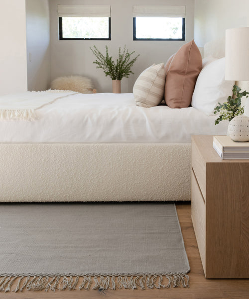 Minimalist bedroom with a white bed, neutral pillows, a wooden nightstand featuring a white lamp and plant, the Hudson Jute Rug for natural texture underfoot, and two small windows letting in natural light.