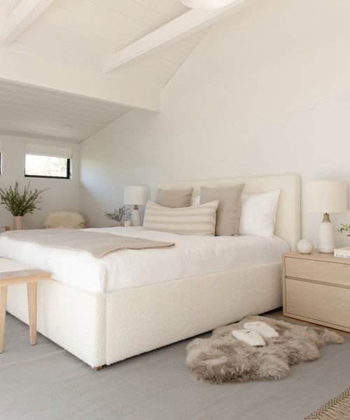 A bright, minimalist bedroom with a white bed, neutral pillows, light wood furniture, soft natural light from small windows, plants, simple table lamps, and a Hudson Jute Rug for a handwoven natural fiber touch.