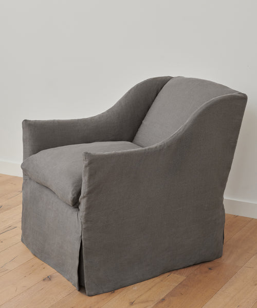 The handmade Miramar Chair, crafted in the USA, features gray Belgian linen with a fitted slipcover, curved arms, and a cushioned seat. It sits on a wooden floor against a plain white wall.