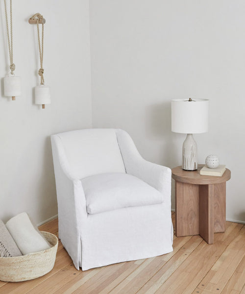 A cozy corner features the white linen Miramar Chair, a wooden side table with a white lamp, a decorative object, and a book. A woven basket with blankets sits on the light wood floor; two hanging decor pieces adorn the wall.