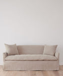 The Miramar Sofa, with its minimalist design and two large Belgian linen cushions, is set against a white wall on a light wooden floor.