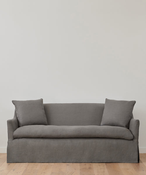 The Miramar Sofa, in simple gray Belgian linen with feather down fill and two matching pillows, sits against a plain white wall on a light wooden floor.