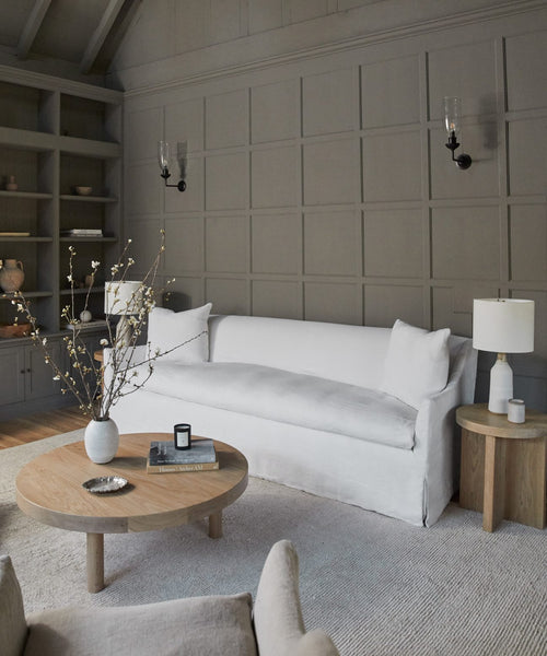 A modern living room with a Miramar Sofa, round wood coffee and side tables, a white lamp, vases with branches, and gray paneled walls featuring built-in shelves and wall sconces.