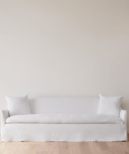 A minimalist room showcases the Miramar Sofa in white Belgian Libeco Linen with two matching pillows, set against a plain light beige wall and light wooden floor.