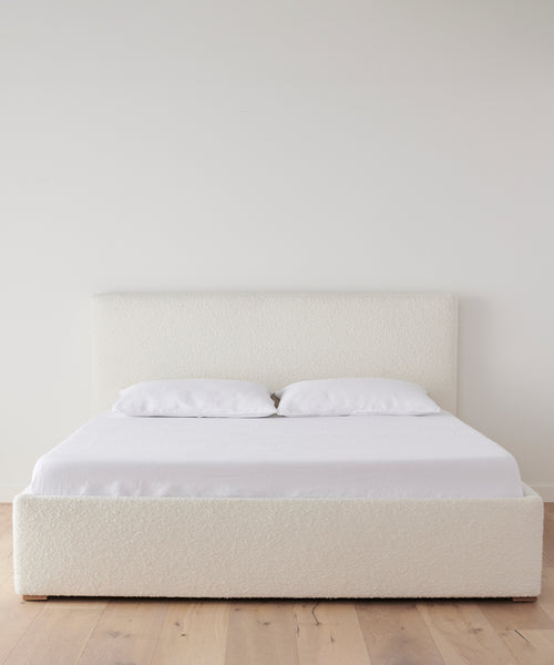 A minimalist bedroom featuring the Pacific Bed in ivory wool boucle with an upholstered headboard, styled with two white pillows and white bedding, set against a plain white wall on a light wooden floor.