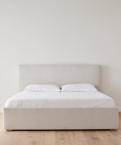 The Pacific Bed features a natural linen frame and upholstered headboard, finished with white pillows and duvet, set against a plain white wall on a wooden floor.