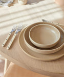 A Pacific Breakfast Bowl from the beige dinnerware collection is placed on a light wooden table, accompanied by two forks and a knife. Sunlight casts gentle shadows across the scene.
