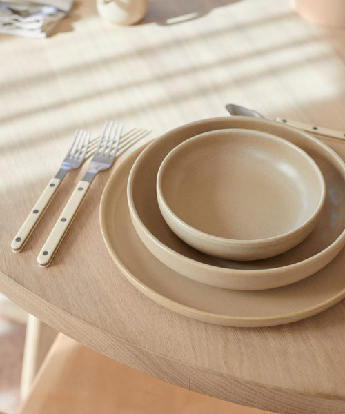 A Pacific Breakfast Bowl from the beige dinnerware collection is placed on a light wooden table, accompanied by two forks and a knife. Sunlight casts gentle shadows across the scene.