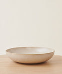 The Pacific Serving Bowl, made of beige stoneware with a shallow, round design, rests on a light wooden surface and off-white background—ideal for upgrading your dinnerware collection.