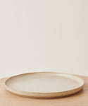 The Pacific Platter, a simple round beige stoneware serving plate with a slightly raised rim, rests empty on a light wooden surface against an off-white background.