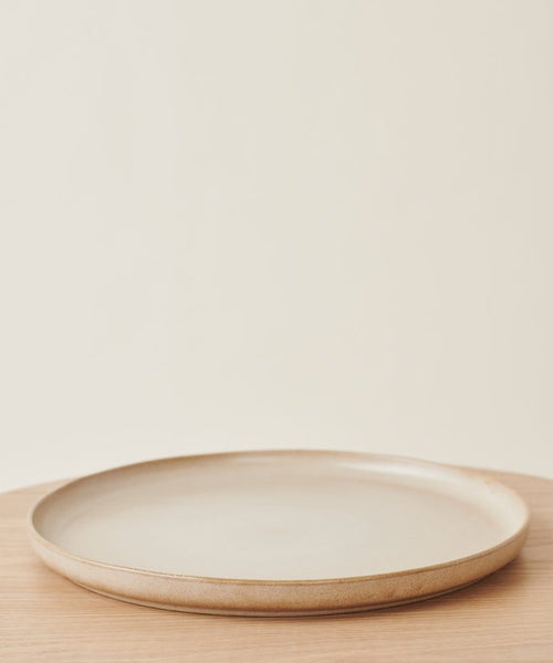 The Pacific Platter, a simple round beige stoneware serving plate with a slightly raised rim, rests empty on a light wooden surface against an off-white background.