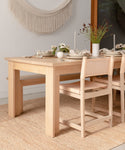 A light wooden dining table with matching chairs is set for a meal atop the Sedona Jute Rug, featuring white plates, glasses, candles, and a vase of flowers. A macrame wall hanging decorates the background.