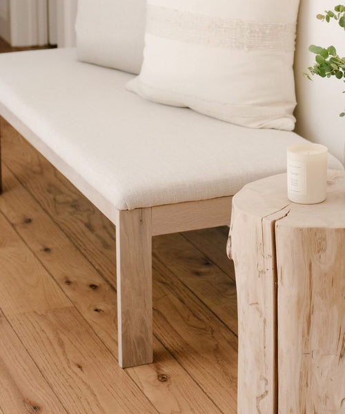 A light wooden bench with a white cushion in Fabric Swatch Textured Natural Linen sits beside a rustic stump table holding a white candle and plant, all atop a light wood floor in a cozy, minimal room inspired by Jenni Kayne Furniture.