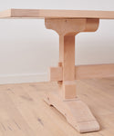 A close-up of the Trestle Dining Table showcases its sturdy handcrafted base and joinery, set on a matching wood floor against a white wall.