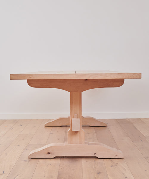 The Trestle Dining Table, handcrafted from Knotty Alder with a central pedestal base, is placed on a light wood floor against a plain white wall and viewed directly from one end.