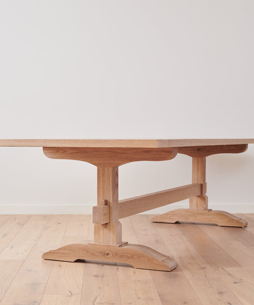 The Trestle Dining Table, handcrafted from oak with a natural finish and sturdy double pedestal legs, sits elegantly on a light wood floor against a plain white wall.