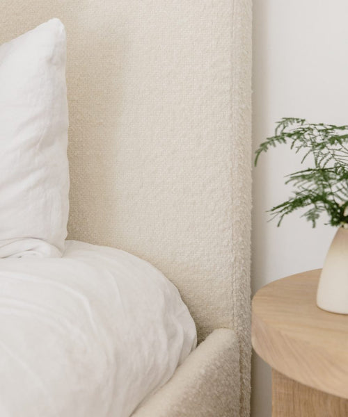 Close-up of a minimalist bedroom with a white bed, beige textured headboard, and wooden nightstand, completed by an upholstered bench in Fabric Swatch Ivory Wool Boucle for a serene touch.
