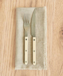 A fork and knife from the Bistro Vintage Finish Cutlery Set, featuring cream handles, rest side by side on a beige linen napkin with frayed edges atop a light wood table.