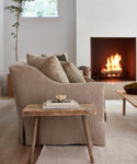 A cozy living room featuring the Miramar Sofa in Belgian linen, a rustic wooden side table with books and a cup, light wood floors, a glowing fireplace, and a window streaming in natural light.