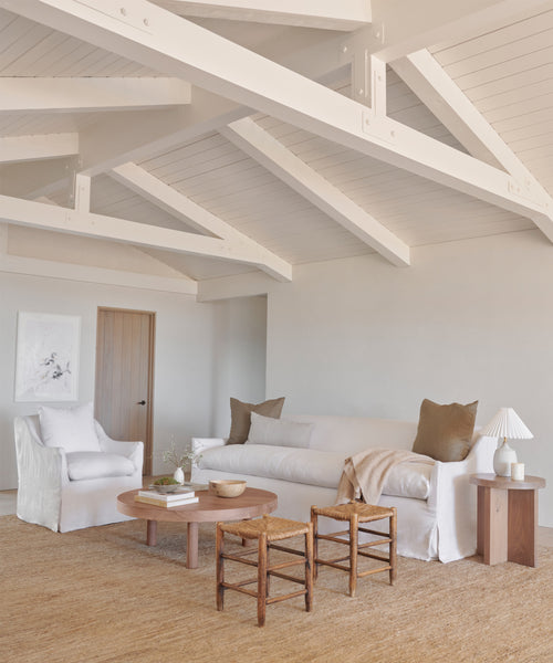 A bright, minimalist living room with white walls and exposed beams showcases a plush Miramar Sofa, an armchair, two wooden stools, a round coffee table on a jute rug, neutral cushions, and a table lamp.