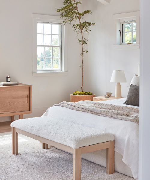 Bright, minimalist bedroom with white walls, a wooden bed and the Otto Bench, light oak furniture, ivory wool boucle bedding, a small table lamp, and a tall potted plant by the window letting in natural light.