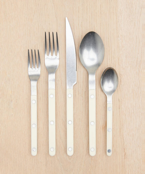 Five utensils—two forks, a knife, a spoon, and a teaspoon—from the Bistro Vintage Finish Cutlery Set with cream handles and silver heads, arranged neatly on a light wooden surface.