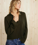 A woman with wavy blonde hair wears the Cashmere Frances Polo in dark olive and blue jeans, standing before a light wooden wall. She looks to the side, one hand touching her collar.