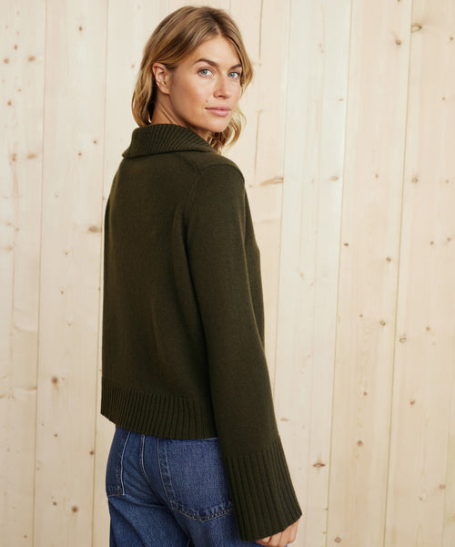 A woman with wavy blonde hair, wearing the Cashmere Frances Polo in dark olive and blue jeans, looks over her shoulder while standing before a light wooden wall.