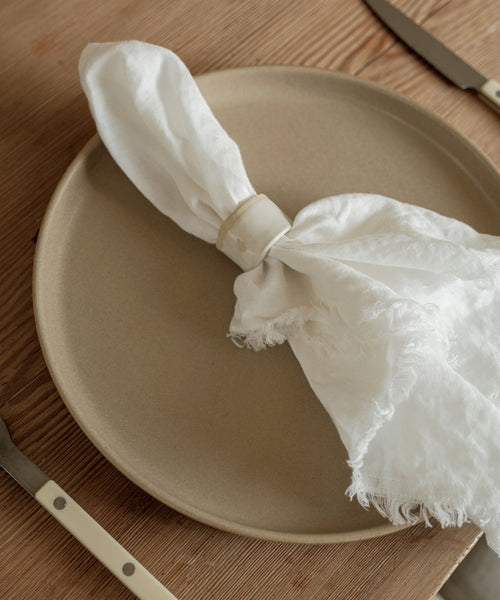 A beige ceramic plate sits on a wooden table with a white napkin in a Ceramic Napkin Rings Set of 4. Cream-handled fork and knife are placed beside the plate.