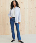 A woman with curly hair stands in front of a light wooden wall, wearing the Boyfriend Shirt in French blue over a white top, paired with blue jeans, a black belt, and black slip-on shoes.