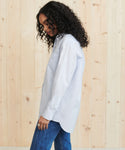 A woman with curly dark hair wears the Boyfriend Shirt in French blue and blue jeans, standing in profile against a light wooden wall.