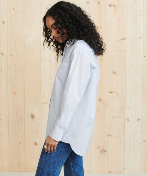 A woman with curly dark hair wears the Boyfriend Shirt in French blue and blue jeans, standing in profile against a light wooden wall.
