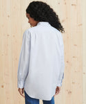 A woman with curly hair, facing away, wears the Boyfriend Shirt in light French blue and blue jeans in front of a light wooden wall.