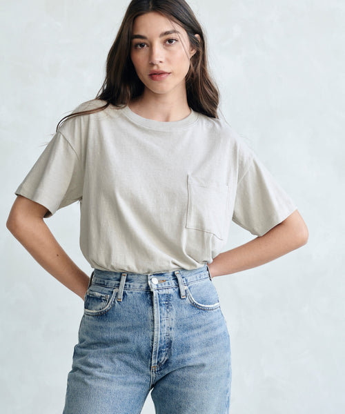 A woman with long brown hair wears the Vintage Pocket Tee in light beige, tucked into high-waisted blue jeans. She stands before a light gray background, hands on hips, highlighting this essential everyday basic.