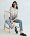 A woman with long brown hair sits casually on a wooden chair, wearing the Vintage Pocket Tee, blue jeans, and black slip-on shoes—an ideal everyday look—set against a light-colored background.