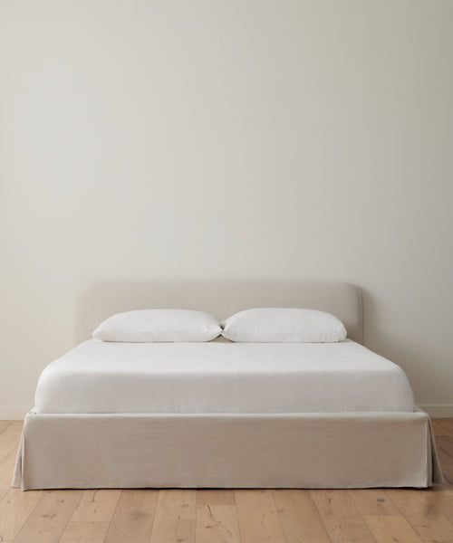 A minimalist bedroom showcases the Cove Bed with a light beige linen headboard, white bedding, and two pillows, set against an off-white wall on a light wood floor.