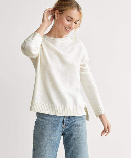 A woman smiles and touches her hair while wearing the Everyday Sweater in ivory with light blue jeans, standing against a plain background.