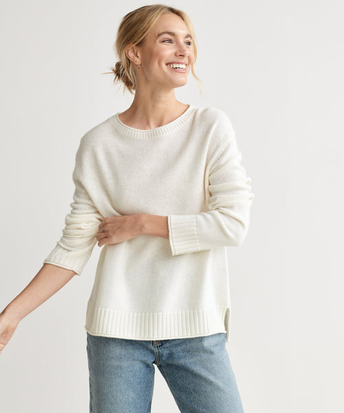 A smiling woman with blonde hair in a loose bun wears the Everyday Sweater in ivory and light blue jeans, standing against a plain white background.