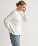 A woman with light brown hair in a low bun wears the Everyday Sweater in ivory and blue jeans, standing sideways with one hand in her pocket and the other touching her forehead against a plain light background.
